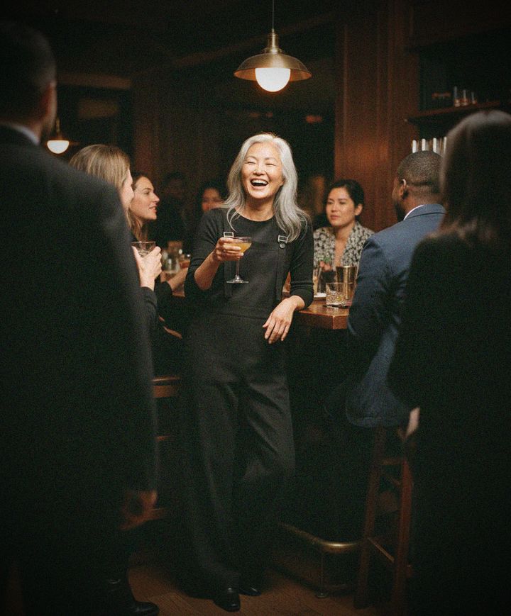A woman with long silver hair laughs at a crowded bar, cocktail in hand, wearing the Deep Slate overall.